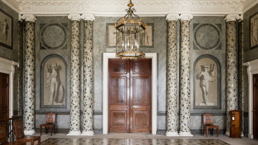 A view of the Entrance Hall at Attingham Park, with double wooden doors, plasterwork ceiling and faux marble pillars visible.